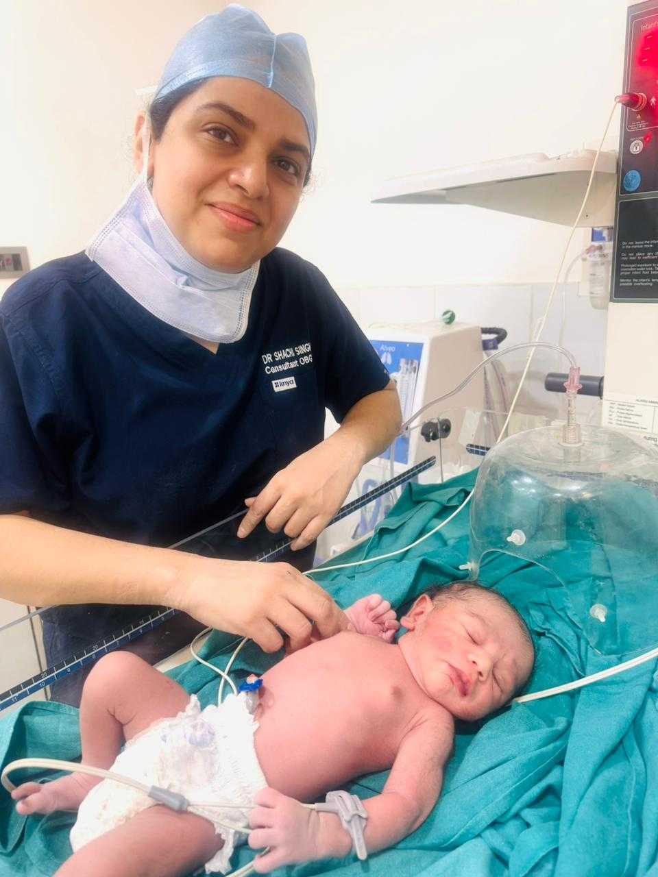 Dr. Shachi Singh in surgical scrubs with a newborn baby on the examination table after delivery